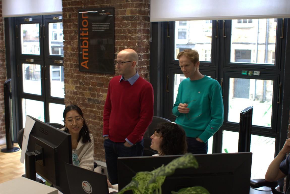 Four professionals in an AI marketing agency collaborating around a desk in a modern office with large windows and an ambition poster