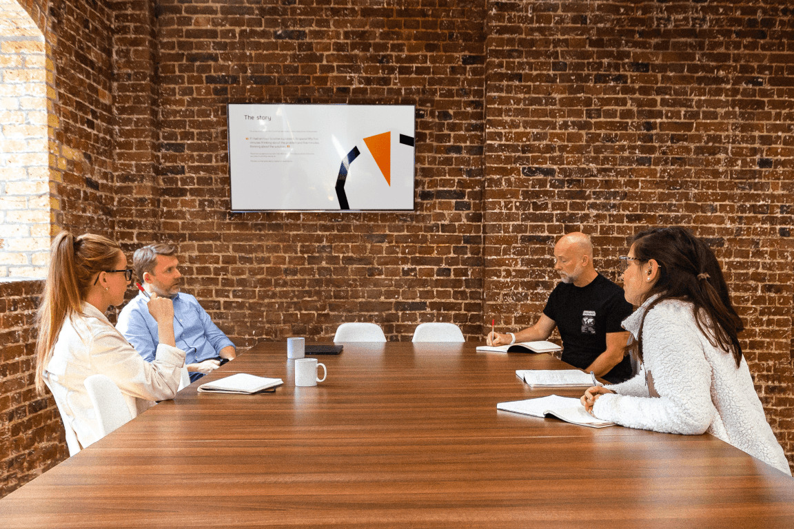 Team participating in a hybrid meeting around a large table in a brick-walled office with remote colleagues on screen